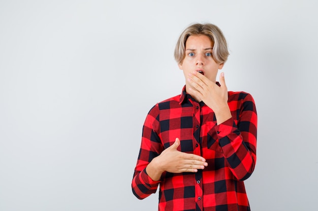 Jeune adolescent en chemise à carreaux gardant la main près de la bouche et l'air terrifié, vue de face.