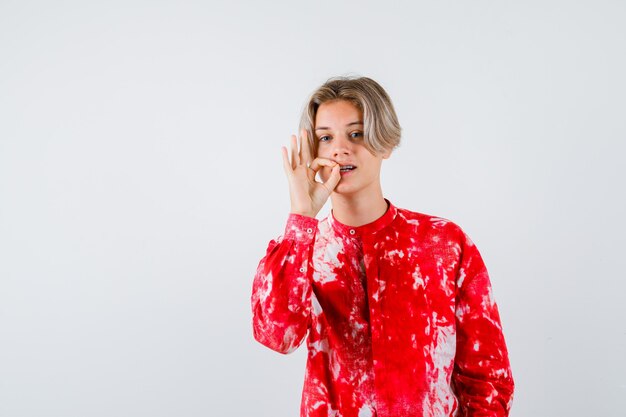 Jeune adolescent en chemise montrant un geste de fermeture éclair et l'air confiant, vue de face.