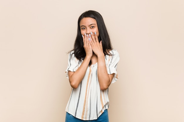 Jeune adolescent chinois mignon Jeune femme blonde portant un manteau contre un mur rose en riant de quelque chose, couvrant la bouche avec les mains.