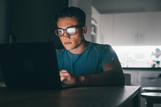 Un jeune adolescent étudie et travaille à la maison la nuit sur la table à l'aide d'un ordinateur portable souriant et s'amusant à faire ses devoirs Lumière de l'écran sur facexA