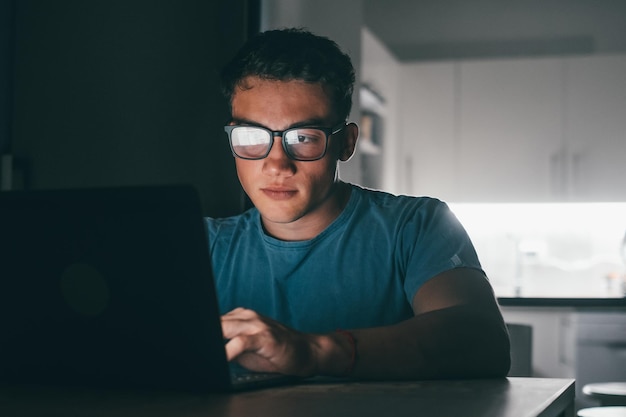 Un jeune adolescent étudie et travaille à la maison la nuit sur la table à l'aide d'un ordinateur portable souriant et s'amusant à faire ses devoirs Lumière de l'écran sur facexA