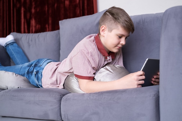 Jeune adolescent garçon à l'aide de tablette allongée sur un canapé gris à la maison