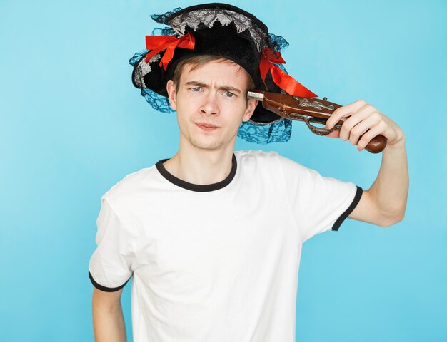 Jeune adolescent mâle drôle en t-shirt blanc sur fond bleu en chapeau de pirate et pistolet à la main veut tirer dans la tête