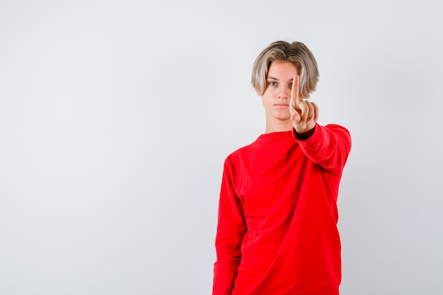 Jeune adolescent montrant un geste de la minute en pull rouge et l'air sérieux, vue de face.