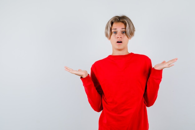 Jeune adolescent en pull rouge montrant un geste impuissant et l'air perplexe, vue de face.