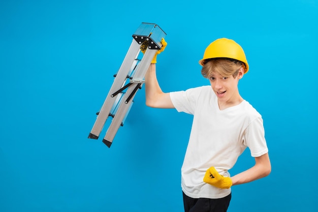 Jeune adolescent réparateur en t-shirt blanc et gants avec un casque jaune dans la tête tenant une échelle dans les mains debout en studio sur fond bleu Un futur architecte isolé sur fond bleu