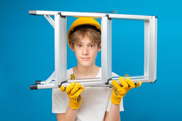 Jeune adolescent réparateur en t-shirt blanc et gants avec un casque jaune dans la tête tenant une échelle dans les mains debout en studio sur fond bleu Un futur architecte isolé sur fond bleu