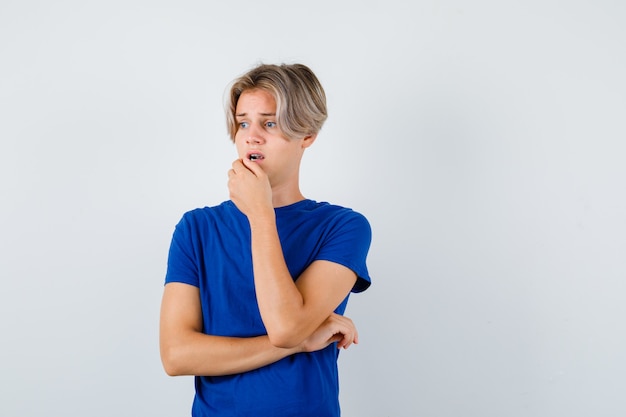 Jeune adolescent en t-shirt bleu avec la main sur le menton tout en détournant les yeux et l'air anxieux, vue de face.