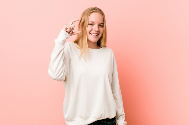 Jeune adolescente blonde tenant quelque chose de petit avec les index, souriant et confiant.