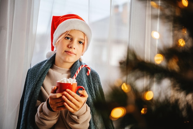 Jeune adolescente en bonnet de noel ayant une tasse de chocolat chaud avec de la guimauve avec un sapin de noel sur t...