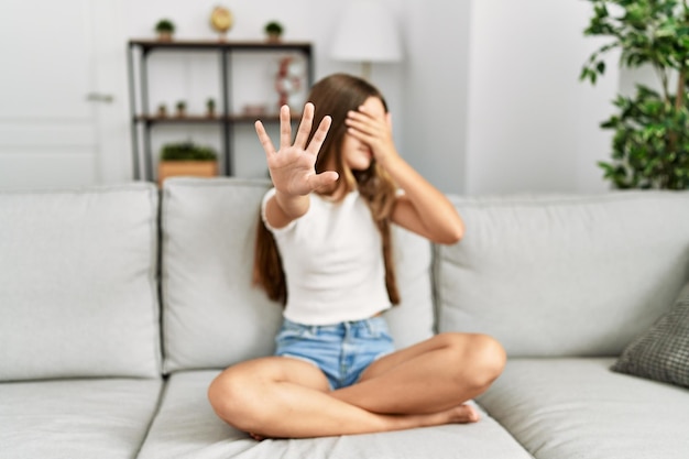 Jeune adolescente brune assise sur le canapé à la maison couvrant les yeux avec les mains et faisant un geste d'arrêt avec une expression de tristesse et de peur. concept embarrassé et négatif.