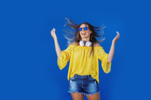 Jeune adolescente caucasienne joyeuse en chemisier jaune et short en jean sautant devant le mur bleu.