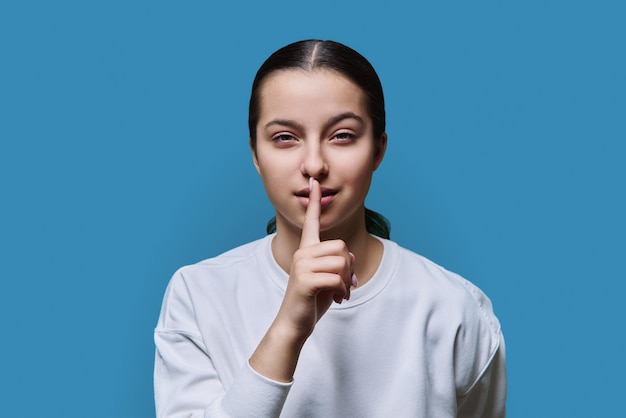 Jeune adolescente montrant un geste silencieux avec le doigt sur les lèvres fond bleu studio Fille étudiante regardant la caméra Langage corporel chut silence chut potins mystère concept