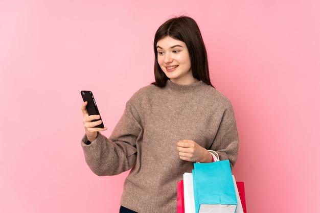 Jeune adolescente sur mur rose tenant des sacs à provisions et écrivant un message avec son téléphone portable à un ami