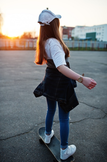 Jeune adolescente urbaine avec planche à roulettes, porter des lunettes, casquette et jeans déchirés sur le terrain de sport.