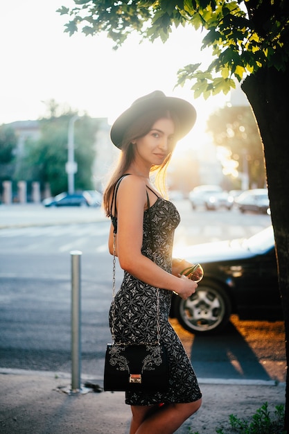 Jeune adolescente vêtue d'une robe blanche et d'un chapeau. style de la grande ville. Ciel coucher de soleil et rayons du soleil. Beauté naturelle jolie femme en chapeau de hipster fedora noir, sur le coucher du soleil chaud du soir d'été dans la grande ville