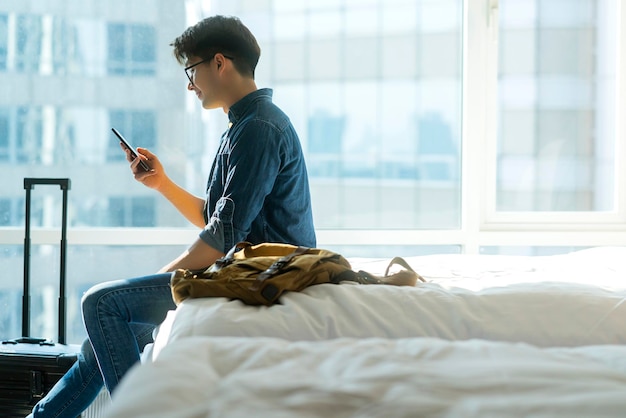 Jeune adulte asiatique voyageur mâle tissu décontracté avec veste en jean main utiliser la conversation sur smartphone sur un lit moelleux blanc avec sac à dos sur le concept de voyage de week-end de vacances