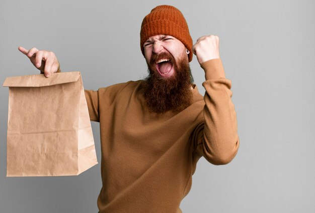 Jeune adulte barbu aux cheveux roux avec un sac en papier avec de la nourriture à emporter