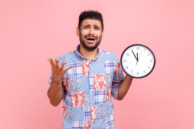 Jeune adulte barbu nerveux mécontent aux cheveux noirs criant, montrant l'horloge murale à l'écran, date limite, portant une chemise bleue de style décontracté. Tourné en studio intérieur isolé sur fond rose.