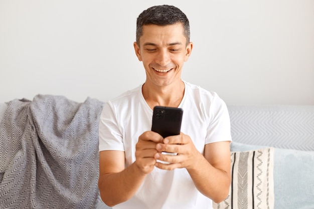 Jeune adulte caucasien optimiste aux cheveux noirs portant un t-shirt blanc assis sur un canapé et utilisant un téléphone intelligent, vérifiant les réseaux sociaux ou tapant des messages, exprimant des émotions positives.