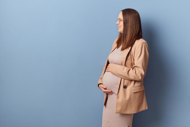 Une jeune adulte enceinte souriante et joyeuse portant une robe et une veste posant isolée sur un fond bleu tenant son ventre en regardant de l'autre côté de l'espace de copie pour le texte promotionnel