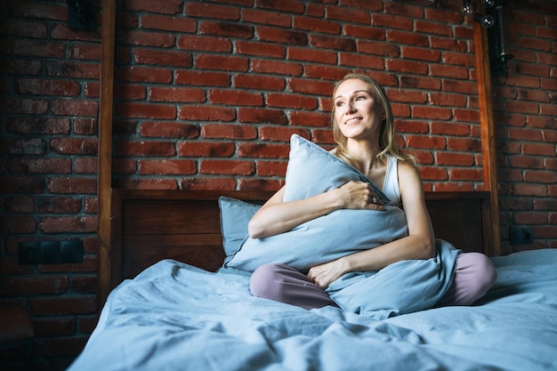 Jeune adulte femme souriante aux cheveux longs blonds en décontracté assis sur le lit avec un oreiller à la maison