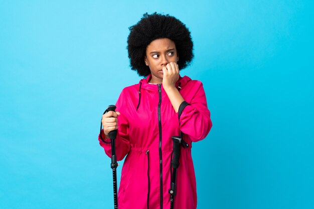 Jeune afro-américain avec sac à dos et bâtons de randonnée isolés sur bleu nerveux et peur de mettre les mains à la bouche