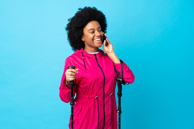 Jeune afro-américain avec sac à dos et bâtons de randonnée sur le mur bleu en gardant une conversation avec le téléphone mobile
