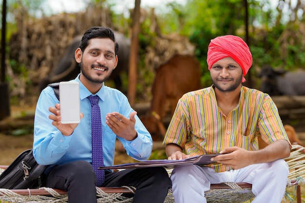 Jeune agent de banque ou agronome indien montrant un smartphone avec un agriculteur dans sa ferme