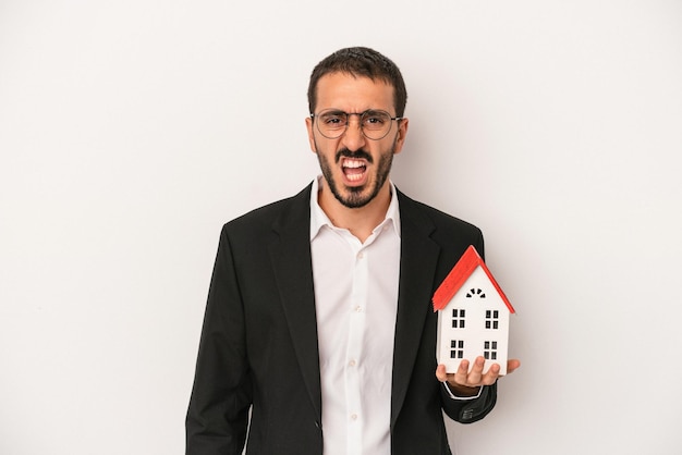 Jeune agent immobilier homme tenant une maison modèle isolée sur fond blanc criant très en colère et agressif