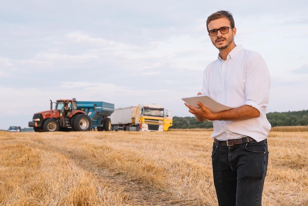 Jeune agriculteur attrayant avec un ordinateur portable debout dans un champ de blé avec une moissonneuse-batteuse en arrière-plan