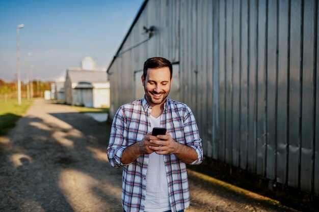 Jeune agriculteur caucasien mal rasé souriant debout à côté de la grange et utilisant un téléphone intelligent pour envoyer des SMS.