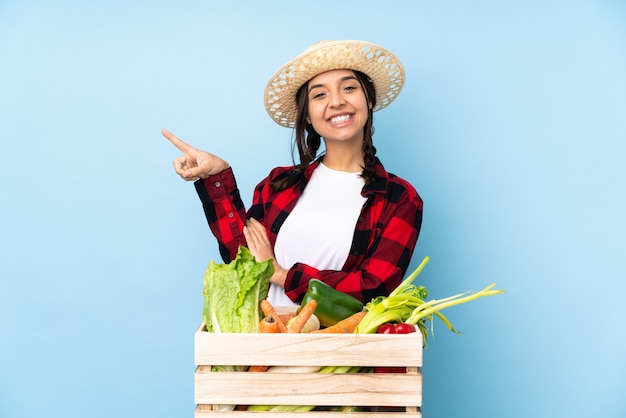 Jeune agriculteur femme tenant des légumes frais dans un panier en bois doigt pointé sur le côté