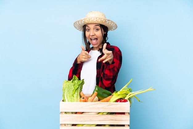 Jeune agriculteur femme tenant des légumes frais dans un panier en bois pointant vers l'avant et souriant