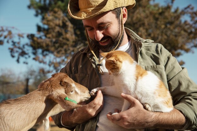 Jeune agriculteur tenant son chat et caressant une chèvre dans sa ferme