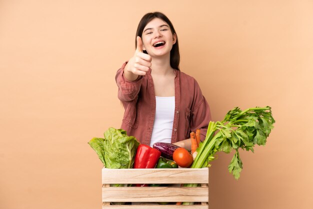 Jeune agricultrice avec des légumes fraîchement cueillis dans une boîte avec les pouces vers le haut parce que quelque chose de bien s'est produit