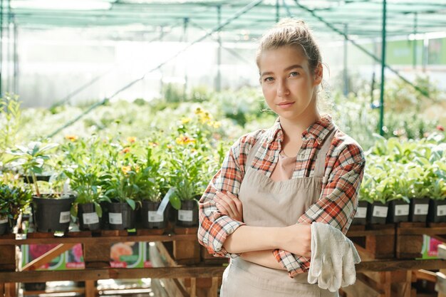 Jeune agricultrice sérieuse avec des gants dans les mains debout et vous regarde contre des plantes vertes dans des pots de fleurs