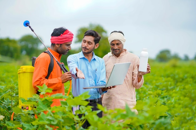 Jeune agronome indien donnant une bouteille d'engrais liquide à un agriculteur et affichant des informations sur le produit dans un ordinateur portable dans un champ d'agriculture verte.