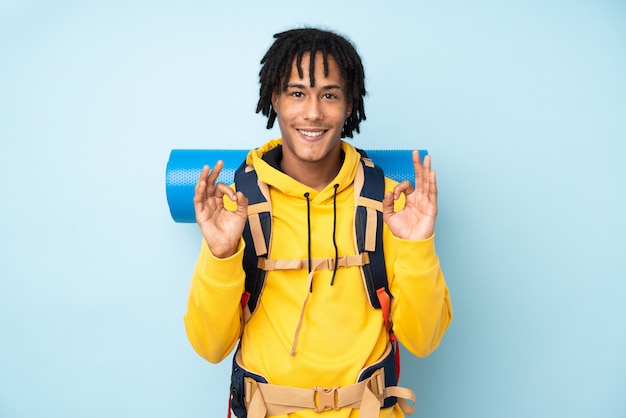 Jeune alpiniste afro-américain avec un gros sac à dos isolé sur fond bleu montrant un signe ok avec les doigts