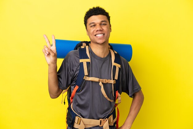 Jeune alpiniste afro-américain avec un gros sac à dos isolé sur fond jaune souriant et montrant le signe de la victoire