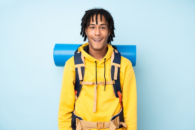 Jeune alpiniste afro-américain avec un gros sac à dos isolé sur un mur bleu avec une expression faciale surprise