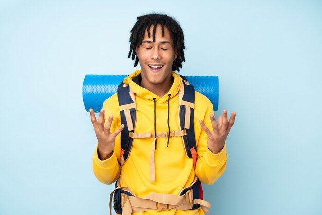 Jeune alpiniste afro-américain avec un gros sac à dos isolé sur un mur bleu malheureux et frustré par quelque chose