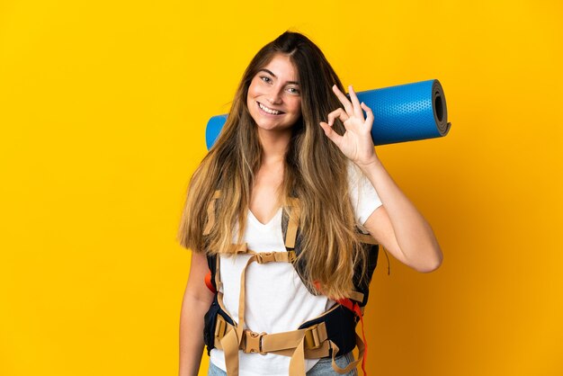 Jeune alpiniste femme avec un gros sac à dos isolé sur mur jaune montrant signe ok avec les doigts