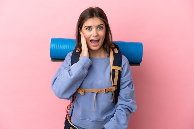 Jeune alpiniste femme avec un gros sac à dos isolé sur un mur rose avec surprise et expression du visage choqué