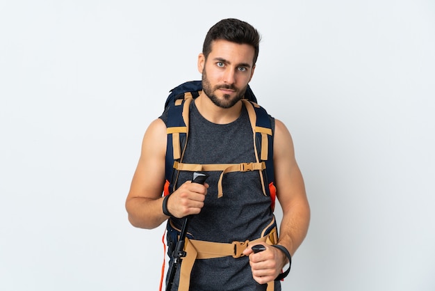 Jeune alpiniste avec un grand sac à dos et bâtons de randonnée isolé sur mur blanc en gardant les bras croisés