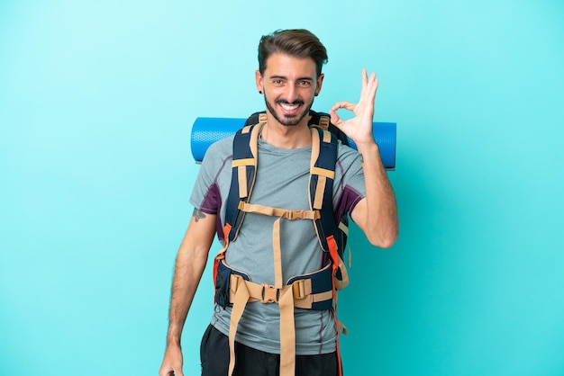 Jeune alpiniste avec un gros sac à dos isolé sur fond bleu montrant un signe ok avec les doigts