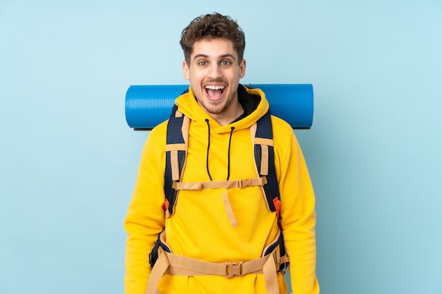 Jeune alpiniste avec un gros sac à dos sur le mur bleu avec une expression faciale surprise