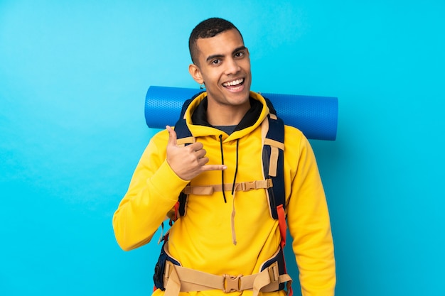 Jeune alpiniste avec un gros sac à dos sur un mur bleu isolé faisant un geste de téléphone