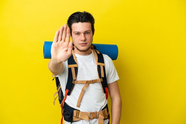 Jeune alpiniste russe avec un gros sac à dos isolé sur fond jaune faisant un geste d'arrêt