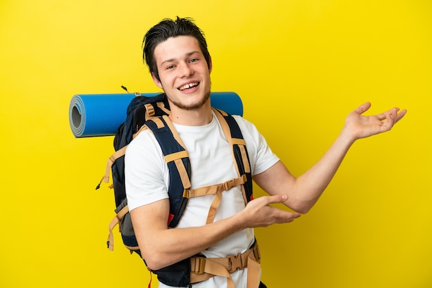 Jeune alpiniste russe avec un gros sac à dos isolé sur fond jaune tendant les mains sur le côté pour inviter à venir